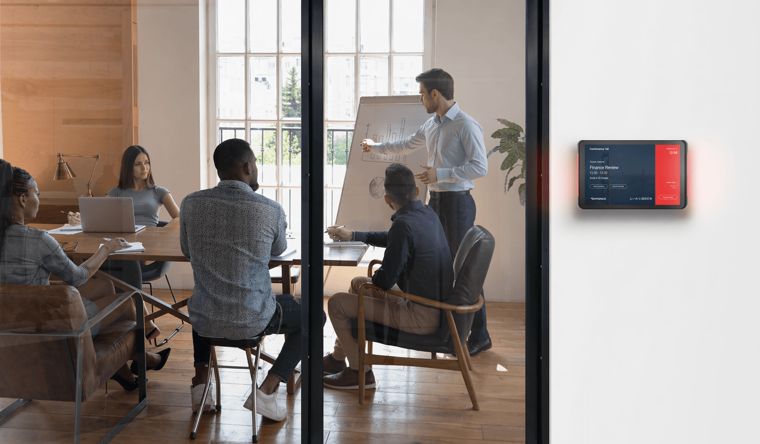 Modern glass-walled meeting room with professionals collaborating, alongside a digital room scheduling display showing real-time meeting information.
